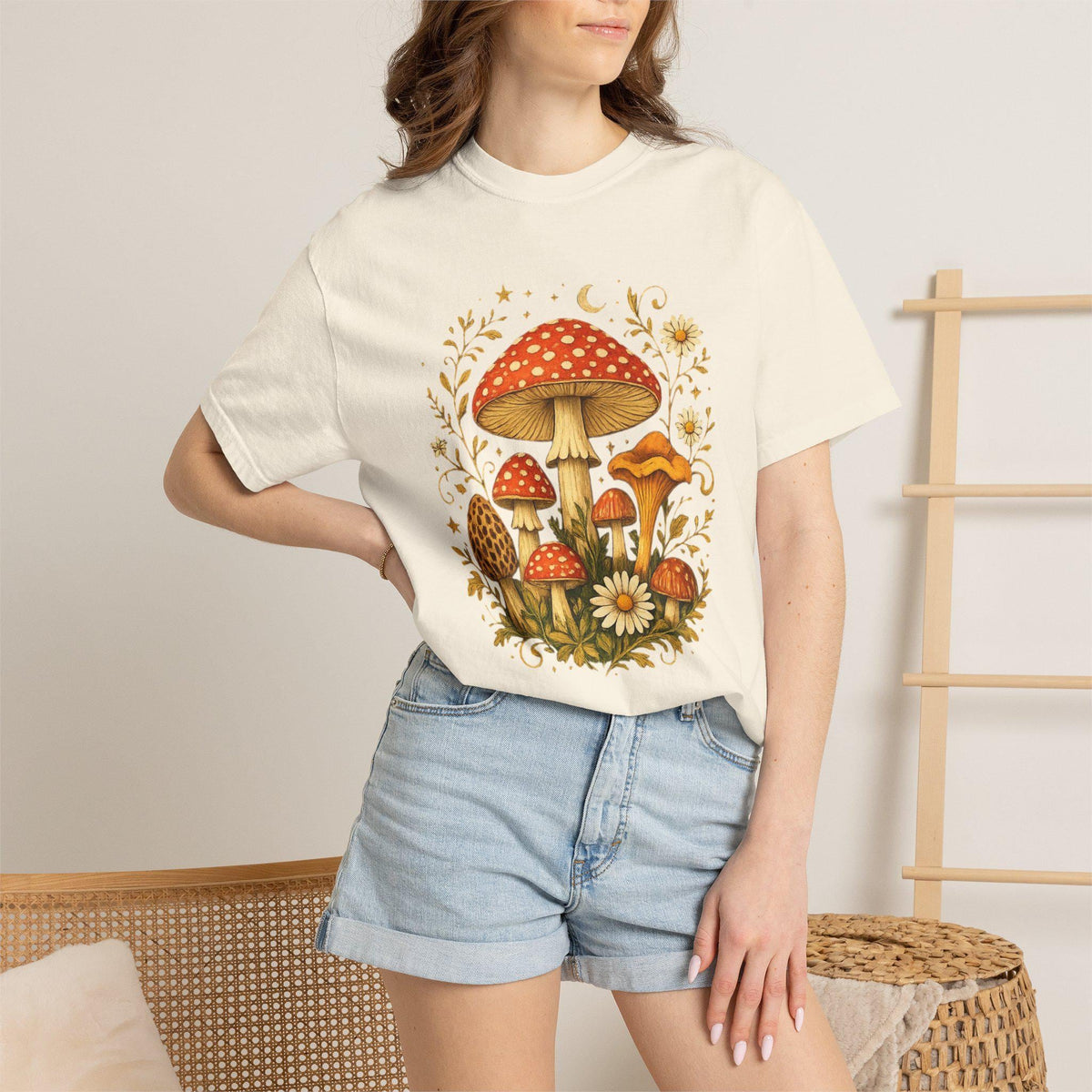 A woman wears an off-white short-sleeved t-shirt with a graphic of red and orange mushrooms, white flowers, and gold leaves, paired with light blue denim shorts.