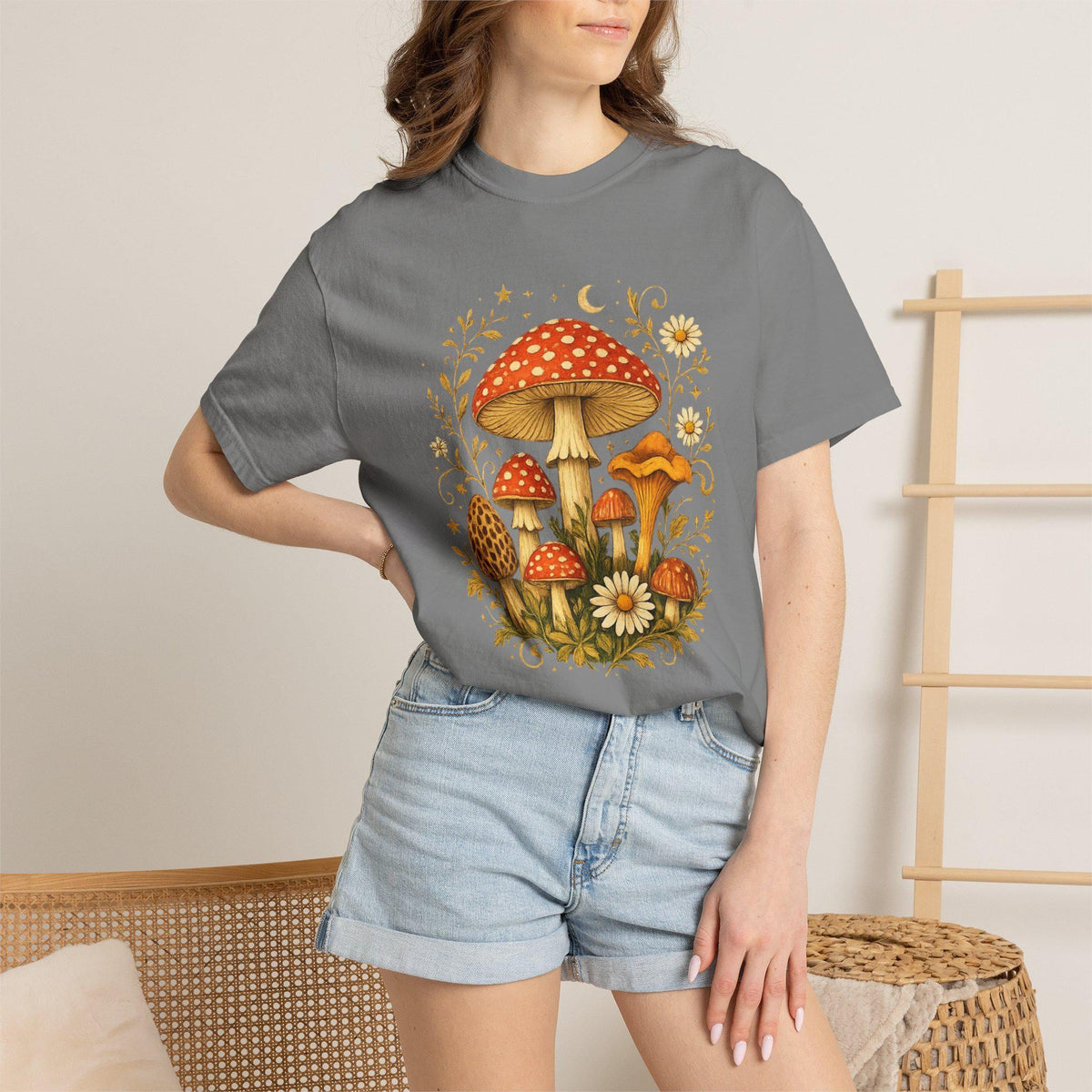 A woman wears a gray short-sleeved t-shirt with a graphic of red and orange mushrooms, white flowers, and gold leaves, paired with light blue denim shorts.