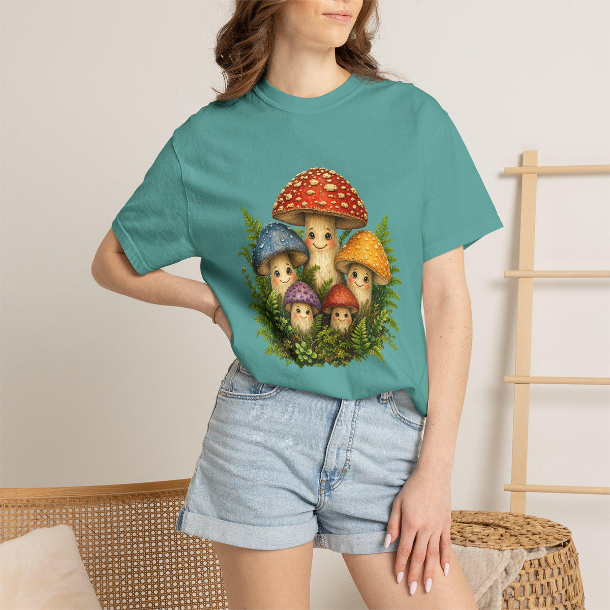 A person wearing a teal t-shirt with a graphic of five smiling mushrooms and green ferns, paired with denim shorts.
