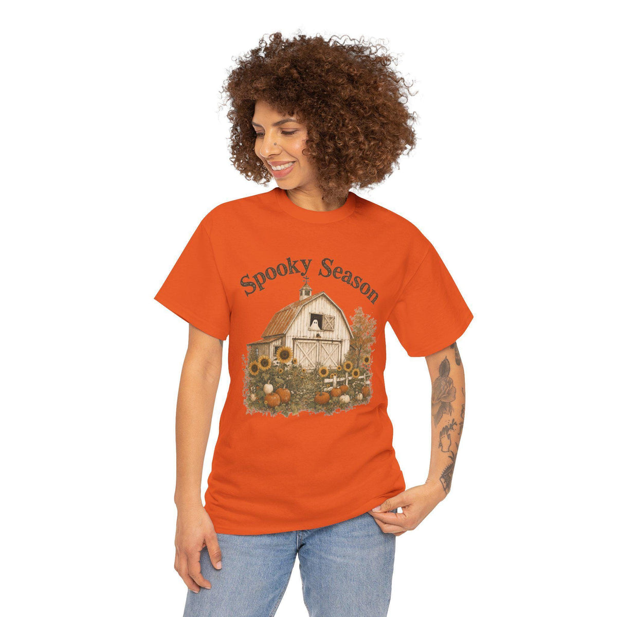 A woman wearing an orange t-shirt with a graphic of a white barn, sunflowers, and pumpkins, with "Spooky Season" text above.
