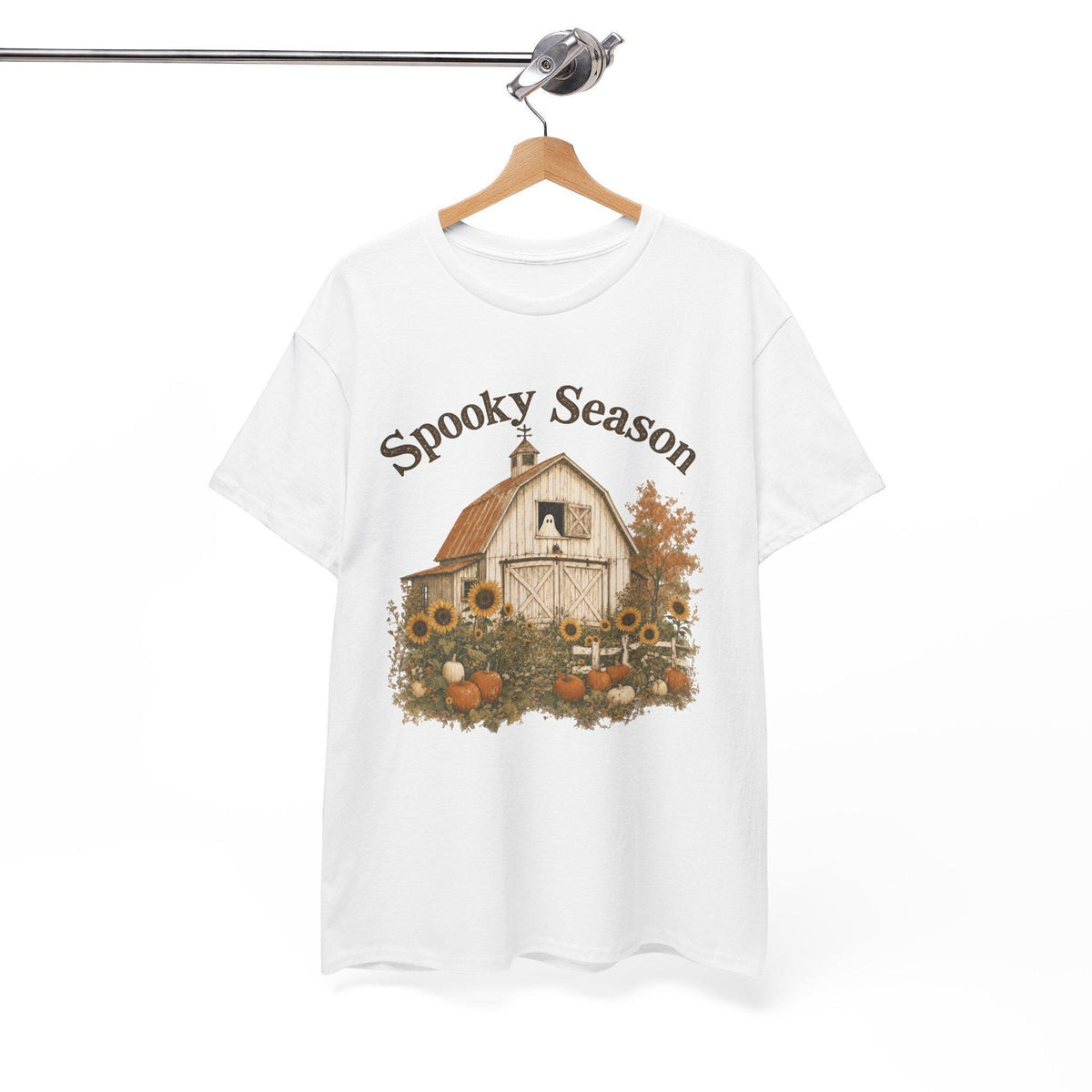 A white t-shirt hanging on a wooden hanger, displaying a graphic of a white barn, sunflowers, and pumpkins, with "Spooky Season" text above.