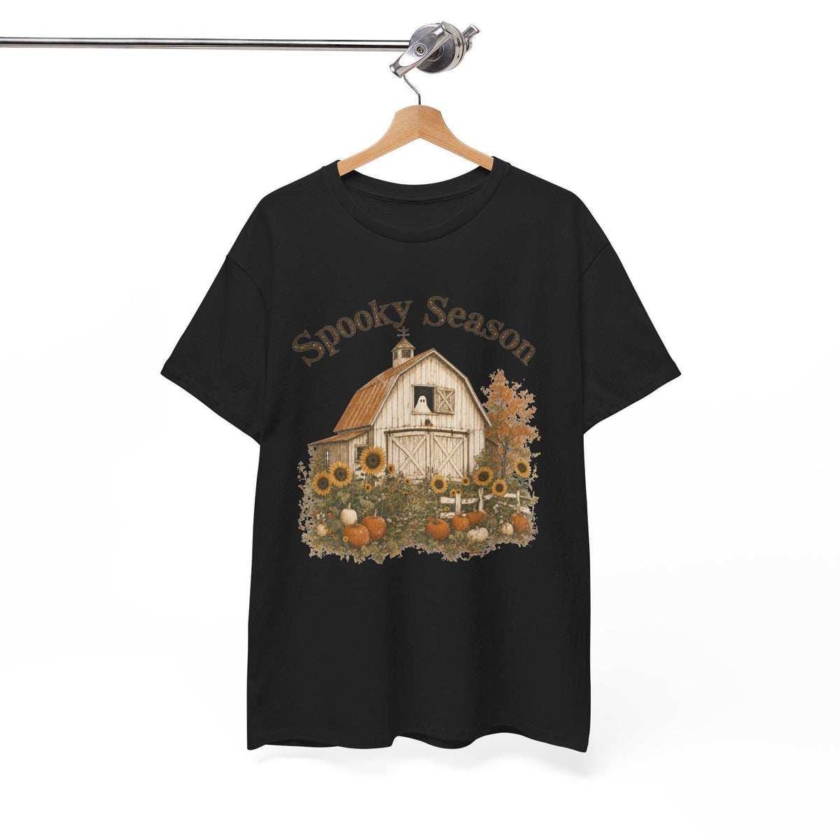 A black t-shirt hanging on a wooden hanger, displaying a graphic of a white barn, sunflowers, and pumpkins, with "Spooky Season" text above.